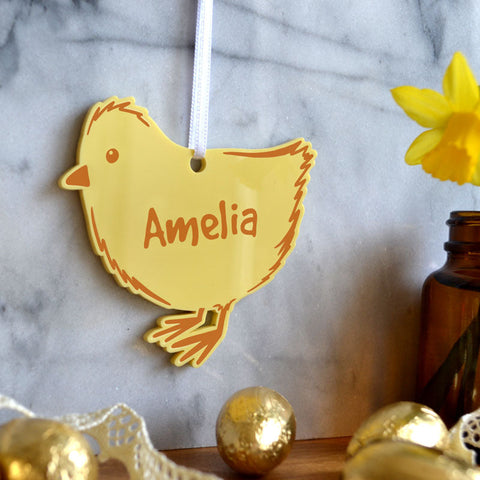 A personalised Easter decoration in the shape of a baby chick. The decoration is made from yellow acrylic and has a name printed in orange in the centre.