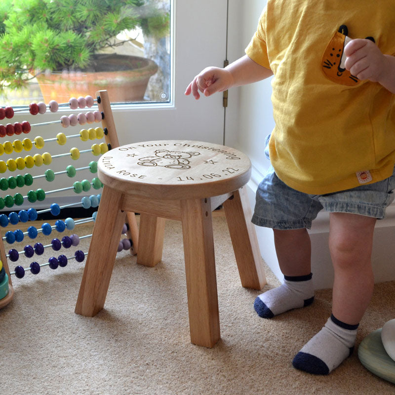 A personalised wooden stool in a children's size. The stool has a design engraved on the wooden seat which features text in a circle around the outside with a teddy bear in the middle.