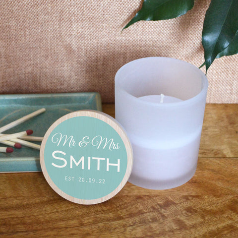 A personalised wedding candle in a glass jar with space for the couple's surname and a date. The design is printed in white lettering on a light blue background..